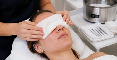 A professional esthetician applying a warm eye compress to a client before a facial extraction treatment to soften pores and improve results.