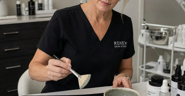 Close-up of a professional esthetician applying a facial mask with a high-quality white fan brush on a client in a spa treatment room.