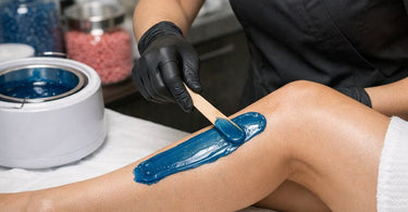 A professional esthetician applying pink hard wax to a client's leg using a wooden spatula, showcasing a stripless waxing technique in a clean, modern spa treatment room.
