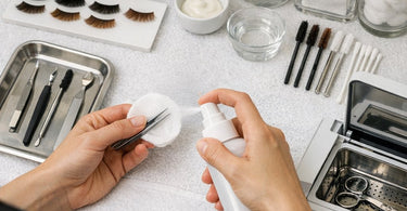 A professional beauty therapist carefully applying lash tint to a client lying down in a spa, with various tint shades visible on a nearby tray.