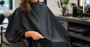 Stack of professionally draped salon capes in various colors on a salon shelf showing different fabric textures and neck closures