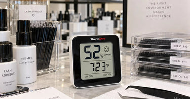 Lash artist checking a digital hygrometer in a professional lash extension treatment room with adhesive tools nearby