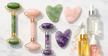 A professional arrangement of a rose quartz Gua Sha tool and a jade facial roller alongside dropper bottles of professional serums on a spa towel