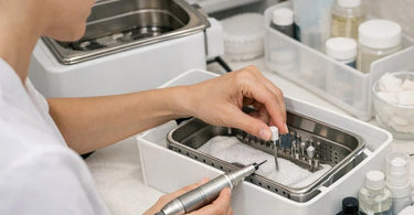 Close up of professional nail technician cleaning an e-file handpiece with a brush and disinfectant spray on a manicure table.