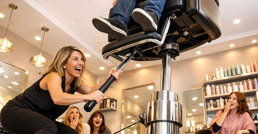 A modern, sleek salon styling chair with black faux leather upholstery and an adjustable hydraulic lift mechanism, shown in a professional salon setting.
