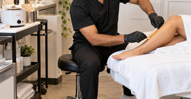 Professional esthetician repositioning a rolling spa stool beside a waxing treatment table for improved working angles and comfortable waxing technique
