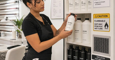 Nail technician safely storing a large gallon jug of acrylic monomer in a cool dark cabinet at a high volume nail salon