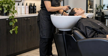A salon stylist standing comfortably on a black anti-fatigue shampoo bowl mat at a backwash station, with a client relaxing in the chair.
