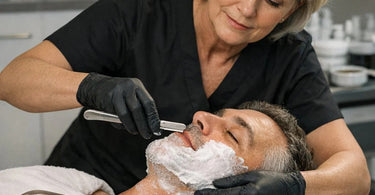 Professional barber holding a straight razor and a selection of stainless steel shaving blades on a marble counter.