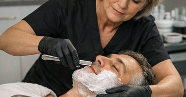 Professional barber holding a straight razor and a selection of stainless steel shaving blades on a marble counter.