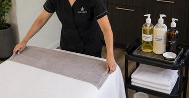 A stack of clean white massage linens and towels neatly arranged, with bottles of massage oil and lotion nearby, symbolizing the battle against oil stains in a professional spa setting.