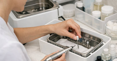 Close up of professional nail technician cleaning an e-file handpiece with a brush and disinfectant spray on a manicure table.