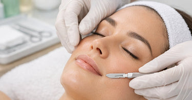 Close-up of a professional esthetician's hands performing safe dermaplaning with a disposable blade on a client's cheek in a clean spa setting.
