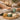 Three glass bowls holding a pink sugar scrub, a creamy white hydrating mask, and a rich yellow foot balm arranged on a spa table with flowers and towels for a pedicure upgrade comparison.