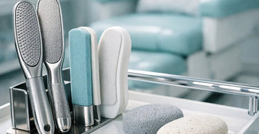 A close up of a professional pedicurist using a stainless steel foot file on a client's heel during a spa pedicure treatment, with various callus removal tools arranged neatly in the foreground.