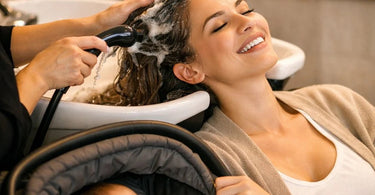 A relaxed new mother smiling as she enjoys a gentle scalp treatment in a salon, with soft lighting and professional tools in view.