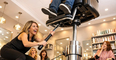 A modern, sleek salon styling chair with black faux leather upholstery and an adjustable hydraulic lift mechanism, shown in a professional salon setting.