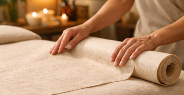 A close-up image of a soft, white biodegradable massage table paper roll being neatly torn along a perforation, laying on a clean massage table.