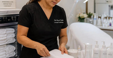 A professional esthetician holds a soft, white facial cleansing towel, ready to use it in a spa treatment, with skincare products in the background.