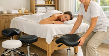 A professional massage therapist sitting comfortably on an adjustable stool next to a massage table, highlighting the importance of ergonomic seating in a spa setting.